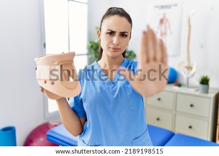 Young physiotherapist woman holding cervical neck collar at medical clinic with open hand doing stop sign with serious and confident expression, defense gesture 