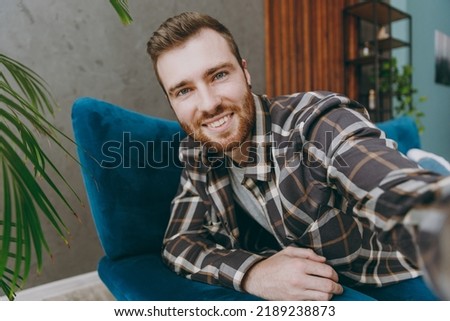 Close up young cool man 20s he wears brown shirt do selfie shot pov on mobile cell phone sitting on blue sofa in own living room apartment stay home indoor flat on weekends. People lifestyle concept