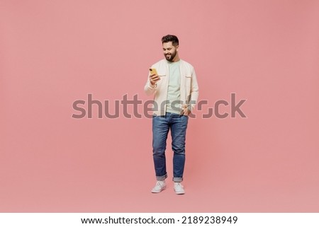 Full body young smiling happy caucasian man 20s wearing trendy jacket shirt hold in hand use mobile cell phone isolated on plain pastel light pink background studio portrait. People lifestyle concept