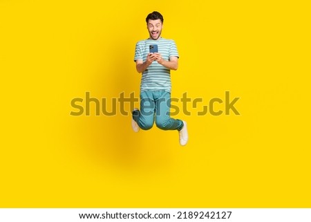 Full size photo of ecstatic impressed guy striped t-shirt blue pants sneakers look on phone jumping isolated on yellow color background