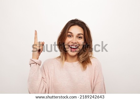 Happy young woman in sweatshirt looking at camera with smile and pointing up while having idea against gray background