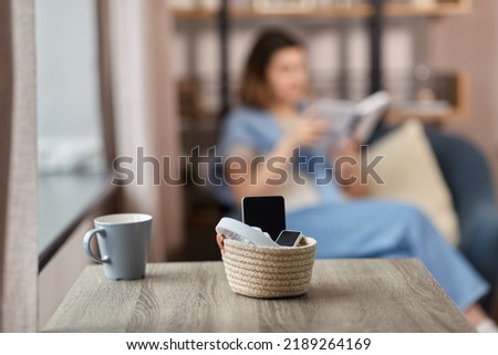 digital detox and leisure concept - close up of gadgets in basket on table and woman reading book at home Royalty-Free Stock Photo #2189264169