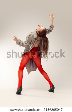 Portrait of young stylish girl posing in red tights and coat posing isolated over grey studio background. Weird dancing moves. Concept of retro fashion, art photography, style, queer, beauty