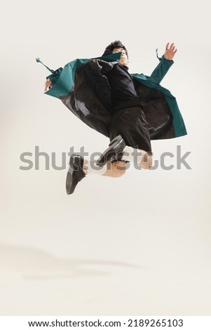 Portrait of stylish young man in black outfit and green coat posing isolated over grey studio background. Jumping . Concept of modern fashion, art photography, style, queer, uniqueness, ad