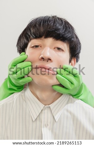 Portrait of young man with piercing posing with hands in rubber gloves on face isolated over grey studio background. Concept of modern fashion, art photography, style, queer, uniqueness, ad