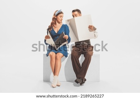 Portrait of young man and woman in retro outfit sitting together, reading magazine or newspaper isolated over white studio background. Concept of retro fashion, style, youth culture, emotions, ad