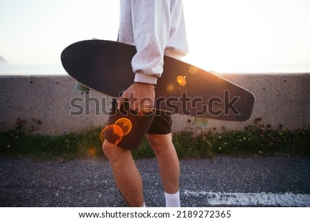 Side view of man carry longboard in his hand. Casual dressed young man in grey hoodie walk with skateboard in sunset light with leaks