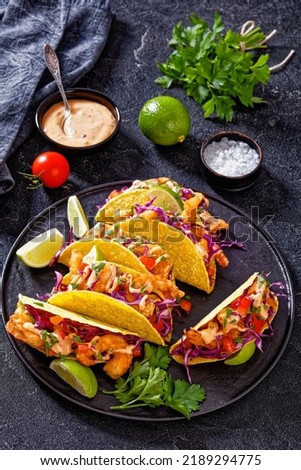 Bang Bang Shrimp Tacos with purple cabbage, tomatoes, parsley, lime and mayonnaise hot chili sauce on black plate on concrete table with ingredients, vertical view from above