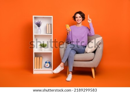 Full length photo of cheerful girl sit chair point finger purchase house interior isolated on orange color background