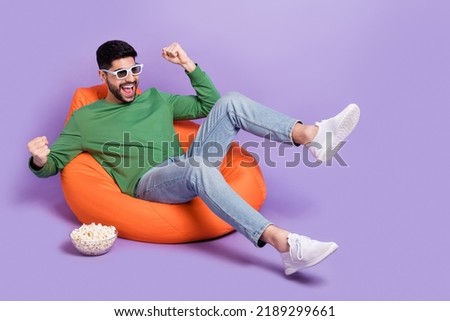 Full body photo of young latin man sit soft beanbag raise fists enjoy movie dressed stylish green look isolated on purple color background