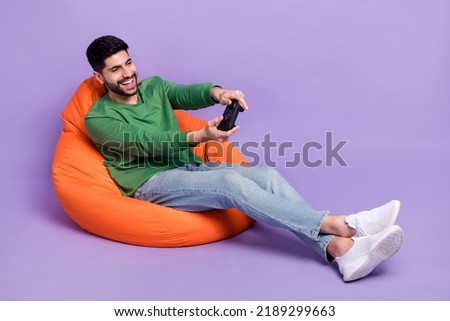 Full size photo of young latin man sitting cozy beanbag hold playstation dressed stylish green garment isolated on purple color background
