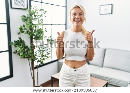 Young caucasian woman standing at living room success sign doing positive gesture with hand, thumbs up smiling and happy. cheerful expression and winner gesture. 