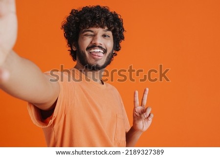 Selfie of young smiling handsome indian man in orange t-shirt blinking and showing victory gesture over isolated orange background