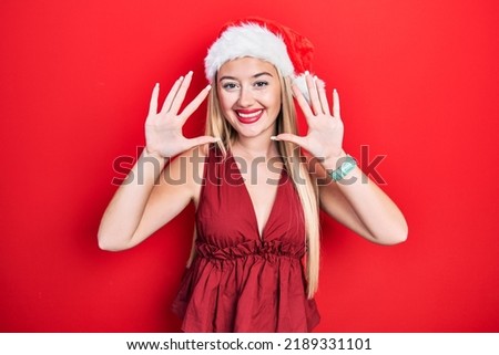 Young blonde girl wearing christmas hat showing and pointing up with fingers number ten while smiling confident and happy. 