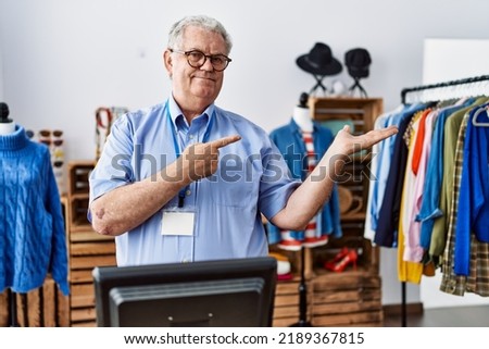 Senior man with grey hair working as manager at retail boutique amazed and smiling to the camera while presenting with hand and pointing with finger. 