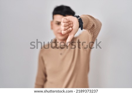 Non binary person standing over isolated background looking unhappy and angry showing rejection and negative with thumbs down gesture. bad expression. 