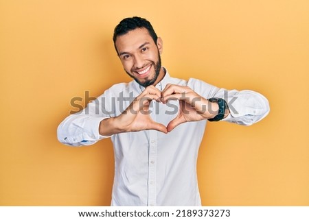 Hispanic man with beard wearing business shirt smiling in love doing heart symbol shape with hands. romantic concept. 
