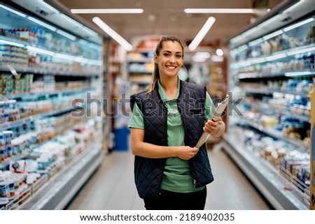 Young happy supermarket manager standing at produce aisle and looking at camera. Royalty-Free Stock Photo #2189402035