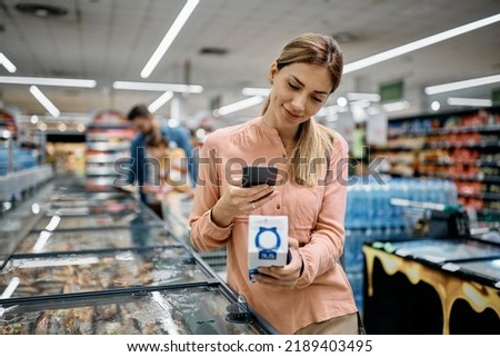 Mid adult woman using smart phone while scanning code on a product in supermarket.  Royalty-Free Stock Photo #2189403495