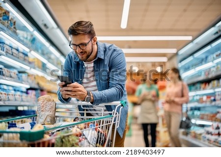 Happy man using mobile phone app while buying groceries in supermarket. 