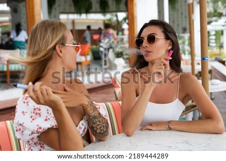 Two young beautiful smiling hipster female in trendy summer sundress.Smoking vapes electronic devices at outdoor cafe. Positive models having fun Royalty-Free Stock Photo #2189444289