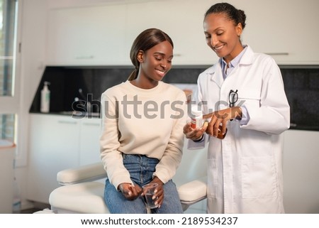 Happy doctor showing vitamins to smiling female patient. Young African American woman sitting at doctors office holding cup of water. Prescription medicine concept Royalty-Free Stock Photo #2189534237