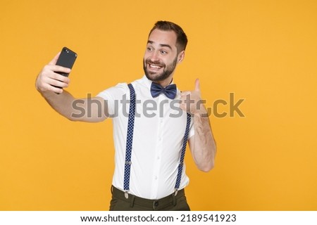 Smiling funny young bearded man 20s in white shirt bow-tie suspender posing doing selfie shot on mobile phone showing thumb up isolated on bright yellow color wall background studio portrait