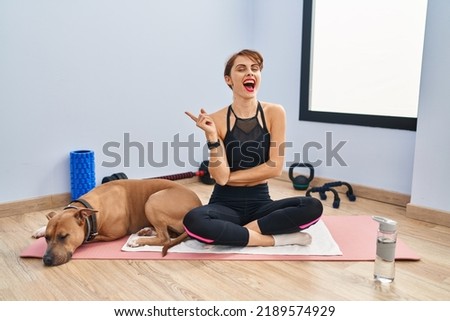 Young beautiful woman sitting on yoga mat with a big smile on face, pointing with hand finger to the side looking at the camera. 