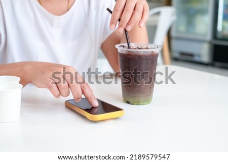 Asian woman in white shirt sitting at white wooden table, holding coffee cup and play smart phone in coffee shop. copy space , Relax time concept