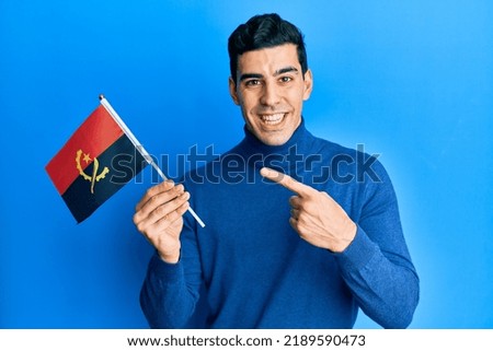 Handsome hispanic man holding angola flag smiling happy pointing with hand and finger 