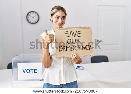 Young blonde woman at political election holding save out democracy banner smiling happy and positive, thumb up doing excellent and approval sign 