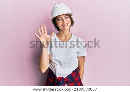 Young caucasian woman wearing hardhat showing and pointing up with fingers number five while smiling confident and happy. 