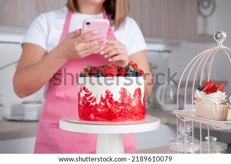 Pastry chef confectioner young caucasian woman takes a picture of the berry-decorated with strawberries and blueberries cake on a stand with her mobile phone. Nearby are cupcakes. Сlose-up