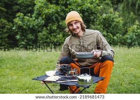 Smiling guy on a hike eats sitting at the table, a man holds a frying pan in his hand, camping in the forest, summer vacation in nature, vacations on foot. High quality photo