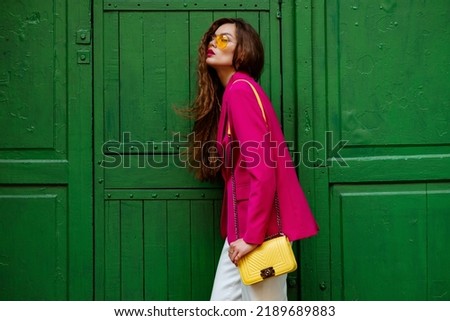 Fashionable confident woman wearing trendy outfit with yellow sunglasses, shoulder bag, pink fuchsia color blazer, posing near green door. Copy, empty space for text Royalty-Free Stock Photo #2189689883