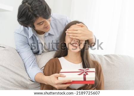 Celebrating anniversary, surprise. Relationship asian young couple love, boyfriend use hand cover girlfriend eye, give a gift by hide box at behind, woman getting present while sitting on sofa at home Royalty-Free Stock Photo #2189725607