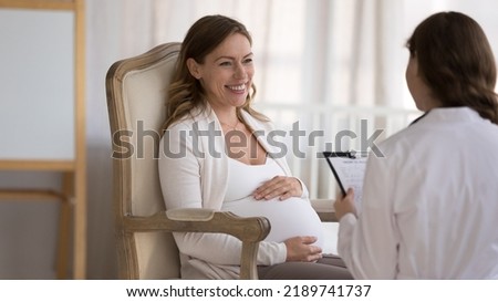 Young smiling pregnant woman patient answers questions of gynecologist during visit at home. Ob-gyn asks about health in late stages of pregnancy, filling medical form, provide prenatal care concept Royalty-Free Stock Photo #2189741737