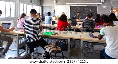 Rear view of group of multiracial high school students in class using laptops while teacher marks exams. Royalty-Free Stock Photo #2189742151