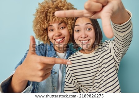 Happy diverse women make frame gesture take good shot measure something on eye picture moment dressed in casual clothes stand closely to each other isolated over blue background. Picturing moment