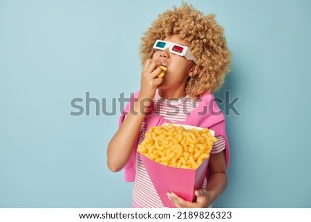 Photo of curly haired woman grabs corn sticks with hunger wears cinema eyeglasses comes in cinema to watch movie dressed in casual clothes spends leisure time with pleasure isolated over blue wall