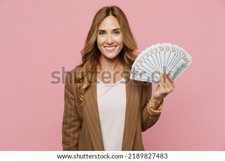 Young fun happy successful employee business woman 30s she wear casual brown classic jacket holding fan of cash money in dollar banknotes isolated on plain pastel light pink background studio portrait Royalty-Free Stock Photo #2189827483