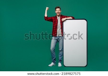 Full body young smiling man he 20s wear red shirt grey t-shirt big huge blank screen mobile cell phone with workspace copy space mockup area do winner gesture isolated on plain dark green background.