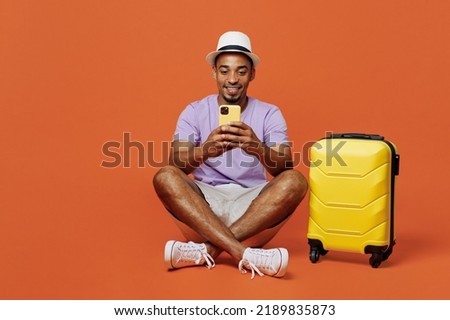 Full body traveler black man wear purple t-shirt hat use mobile cell phone isolated on plain orange color background. Tourist travel abroad in spare time rest getaway. Air flight trip journey concept