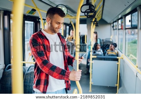 Young cheerful man using a smartphone during his ride and holding onto the bar while standing in a bus. Handsome man taking bus to work while browsing social media. Urban public transportation concept Royalty-Free Stock Photo #2189852031