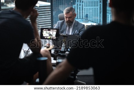 Behind the scenes - Film crew shooting a movie or a corporate video. Adult grey-haired Caucasian CEO businessman actor sitting at in the office located in a skyscraper