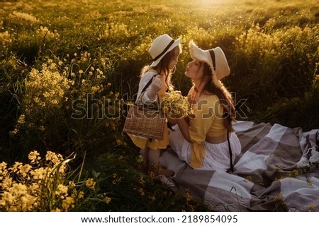 Charming mother and daughter in a white dress and straw hats in a field of yellow flowers at sunset. Family walks, love, motherhood.