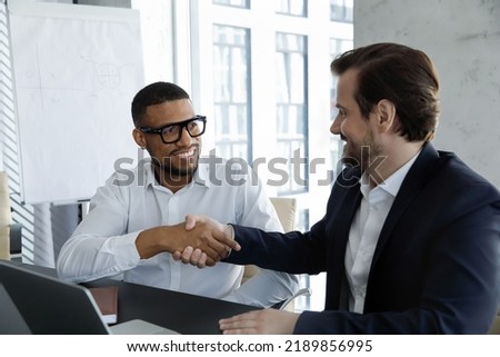 Young African and Caucasian businessmen handshaking accomplish meeting feel satisfied. Multi racial colleagues sit at desk shaking hands express respect and thanking for successful collaborative Royalty-Free Stock Photo #2189856995