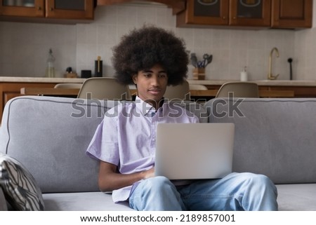 Serious African curly-haired guy sit on sofa with laptop, chatting in social media, enjoy remote communication, share message through messenger app. Young gen use modern tech, blogging, e-date