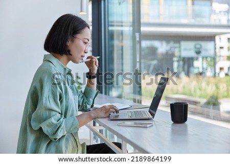 Millennial Asian student prepares for test or exam browses laptop studies in coffee house makes notes in notepad watches online webinar learns foreign language. E learning and online studying Royalty-Free Stock Photo #2189864199