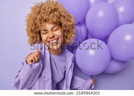Happy carefree woman with curly blonde hair grins at camera keeps eyes closed dressed in stylish jacket celebrates special occasion holds bunch of inflated helium balloons isolated over purple wall Royalty-Free Stock Photo #2189864201